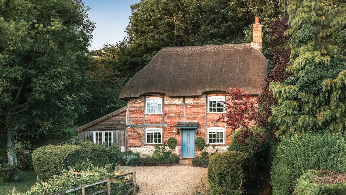 Country, red brick cottage with blue front door, surrounded by trees 